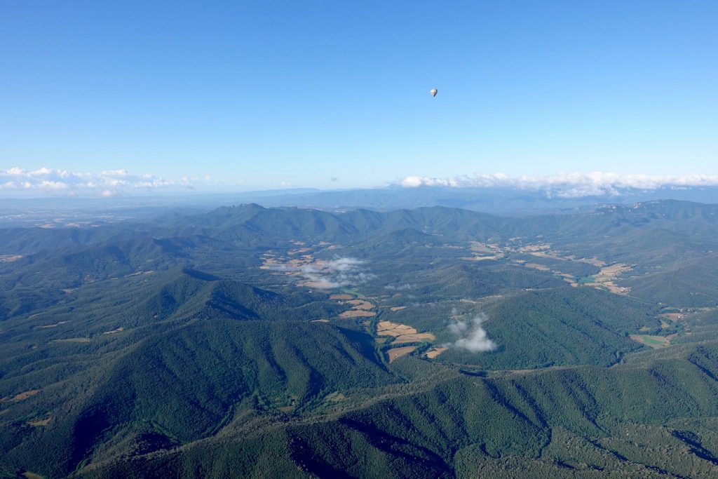 Blick auf die grün bewachsene, hügelige Vulkanlandschaft, am blauen Himmel fliegt in der Ferne ein Heißluftballon