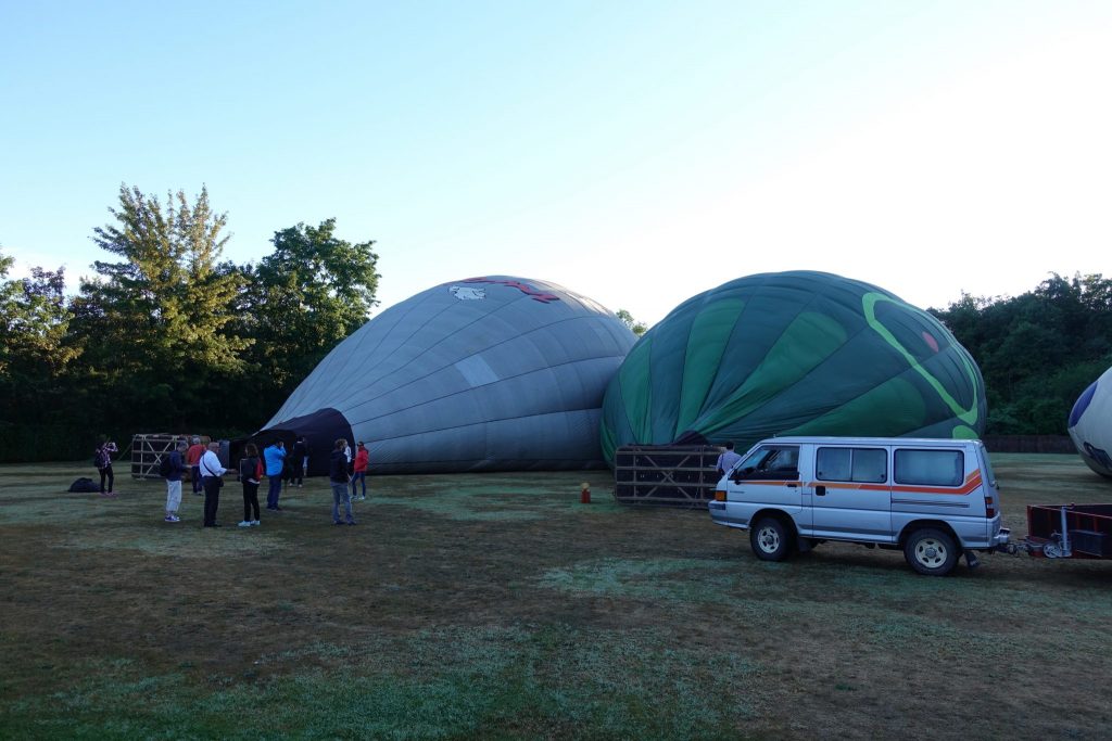 Zwei Heißluftballone liegen am Boden und werden gerade mit Luft gefüllt; rechts im Bild steht ein Auto