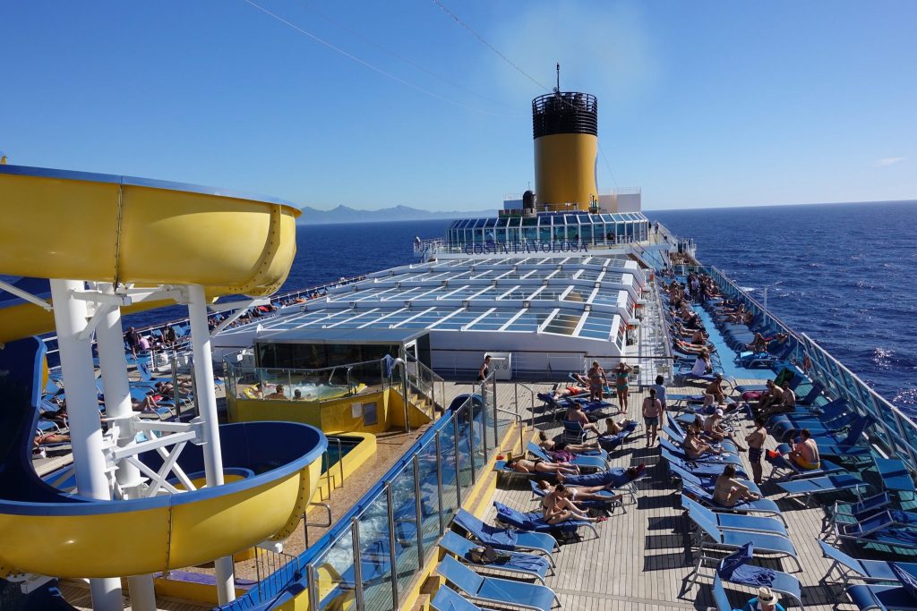 Eine gelbe Rutsche ist links im Bild zu sehen. Das Deck des Kreuzfahrtschiffes ist voll von Liegestühlen