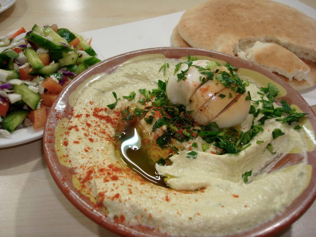 Hummus mit einem harten Ei, bestreut mit Petersilie auf einem Teller im Vordergrund, rechts hinten liegt ein Fladenbrot, links ist ein Teller mit Gemüse