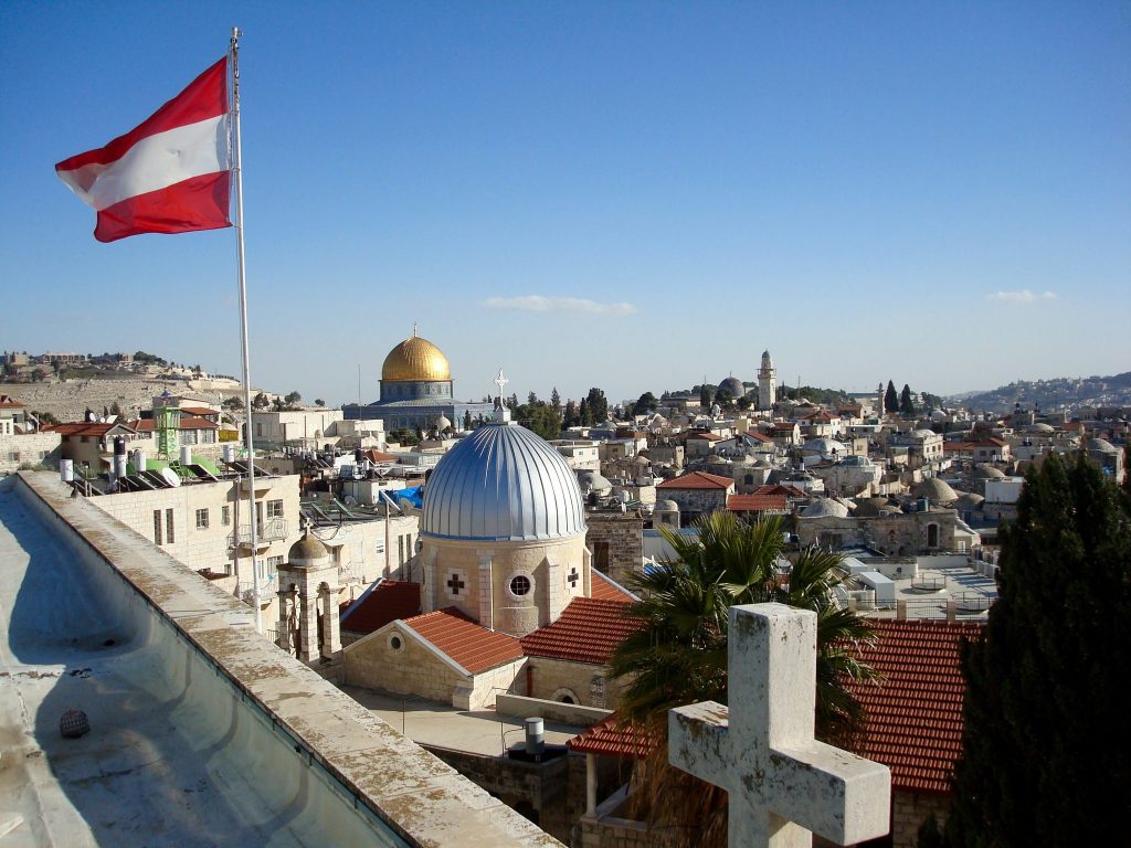 Am Dach des Österreichischen Hospiz in Jerusalem: links weht eine Österreichfahne im Wind, zwei Kuppeln sind zu sehen, vorne rechts ist ein Kreuz